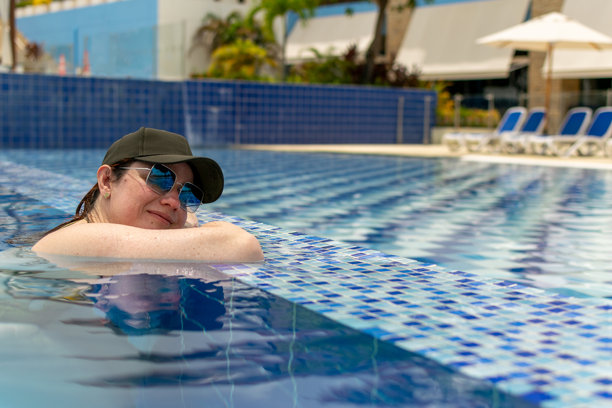 一位戴着黑色棒球帽和太阳镜的宁静女性在碧绿色的泳池中放松。这张图片捕捉了独自旅行中的宁静时刻，反映出一种平和而宁静的度假生活方式。图片下载