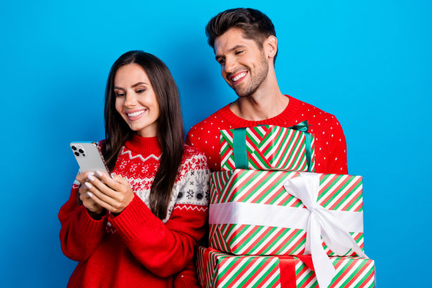 年轻情侣的照片肖像，穿着红色圣诞毛衣，手持电子商务礼物，背景为蓝色。图片下载