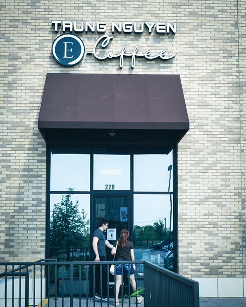 顾客走进位于达拉斯郊区的Trung Nguyen E-Coffee，外观为砖砌立面，醒目的白色标识，圆形E标志，设有坡道入口，展现越南品牌形象。图片下载