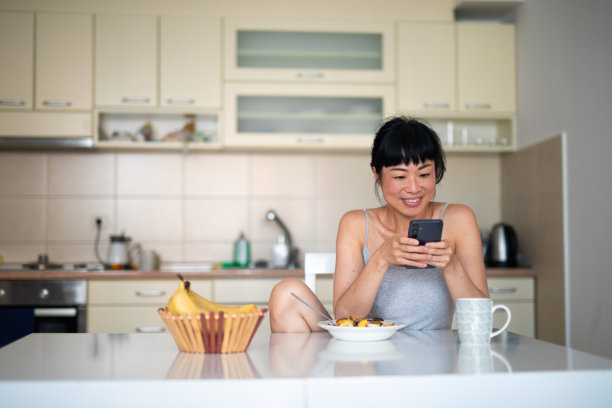 年轻的亚洲女性在家早餐时微笑着使用智能手机 - 股票照片图片下载
