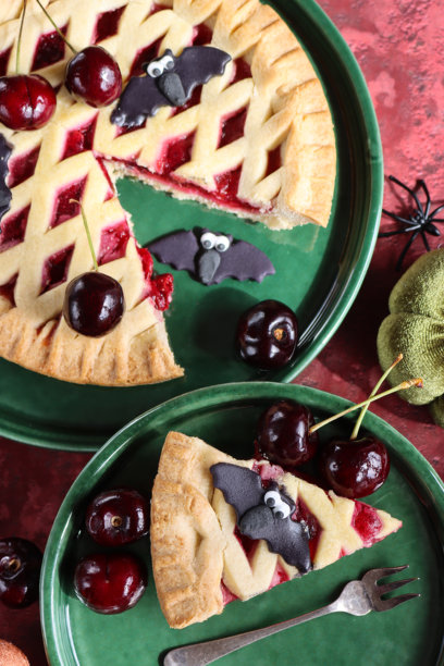 万圣节派对食物的图片，绿色盘子上盛放着新鲜出炉的樱桃挞，金色格子酥皮顶部装饰着黑色蝙蝠翻糖糖霜，切好的派块上满是樱桃，旁边有一个毛绒南瓜，背景为斑驳的红色，俯视角度。图片下载