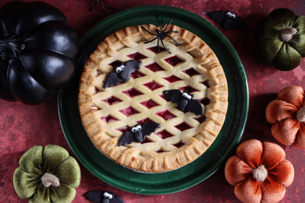 万圣节派对食物的图片，绿色盘子上摆放着新鲜出炉的整颗樱桃挞，金色格子酥皮顶部装饰着黑色蝙蝠翻糖糖饰，塑料蜘蛛、毛绒玩具和瓷制南瓜，斑驳的红色背景，俯视角度。图片下载