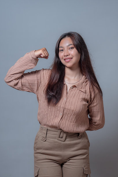 一位自信的印度尼西亚女性，身穿米色衬衫和裤子，微笑着展示肱二头肌，彰显力量。照片在纯灰色背景下捕捉到赋权的神情。图片下载