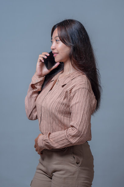 一位印尼女性正在接听电话，她微笑着，一边拿着智能手机一边交谈。她穿着米色的衬衫和裤子，这张照片捕捉了通过科技实现的现代连接。图片下载