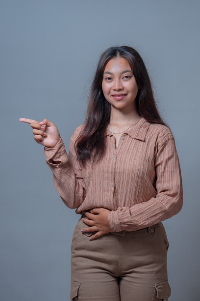 一位印尼女性身穿米色衬衫，一只手叉腰，另一只手向前指向，面带微笑。这是一张在灰色背景下的影棚拍摄照片，展现出友好而自信的风采。图片下载