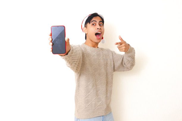 戴着圣诞帽的年轻亚洲男性兴奋地指向空白的智能手机模型屏幕，用于促销和广告，背景为白色，隔离。此图片可用于推广和广告，展示空白屏幕，方便用户添加自己的内容。此图片非常适合用于圣诞节促销、广告活动或任何需要突出智能手机或移动应用程序的场合。此图片非常适合用于圣诞节促销、广告活动或任何需要突出智能手机或移动应用程序的场合。图片下载