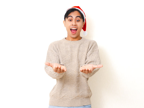 快乐的年轻男子戴着圣诞帽，兴奋地展示双手进行产品推广，背景为白色，用于广告宣传。该图片适合用于广告概念，展示了产品推广和节日气氛。图片下载