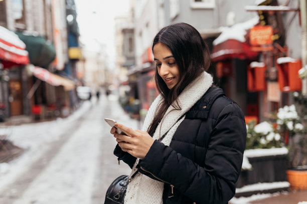 一名年轻女子穿着暖和的衣服，在冬天的雪街市上，一边看着手机里的度假照片，一边微笑着观光。图片下载