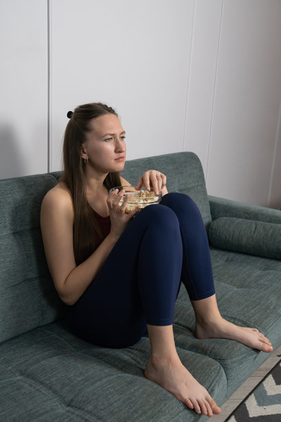 年轻女子坐在沙发上，从玻璃碗里吃着爆米花，全神贯注地观看一部引人入胜的电影，沉浸在屏幕上 unfolding 的情节中。竖图。图片下载
