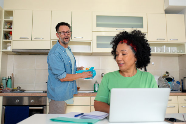 夫妇在厨房多任务处理，男士在洗碗，女士在使用笔记本电脑 - 库存照片图片下载