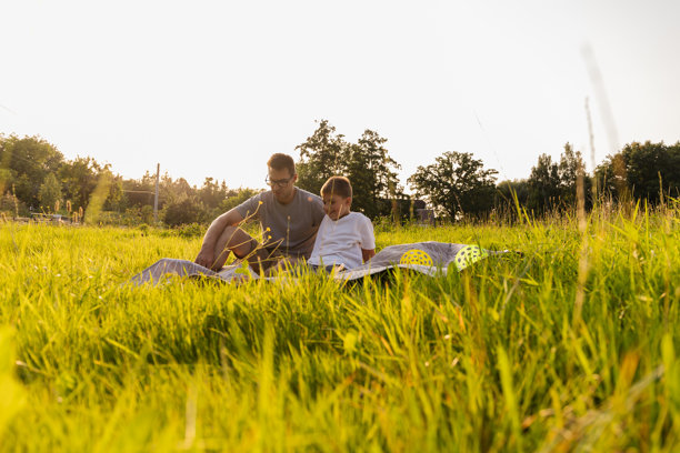 父子俩坐在阳光明媚的草地上，铺着野餐垫，共享温馨时光，享受大自然的美丽。该图片描绘了一个温暖的午后，父子俩在草地上依偎在一起，分享着一段特别的亲子时光，享受着大自然的美丽。他们一起坐在野餐垫上，周围是高高的青草，阳光洒满大地，营造出一种宁静而温馨的氛围。这个画面充满了爱与亲情，展现了人与自然的和谐共处。图片下载