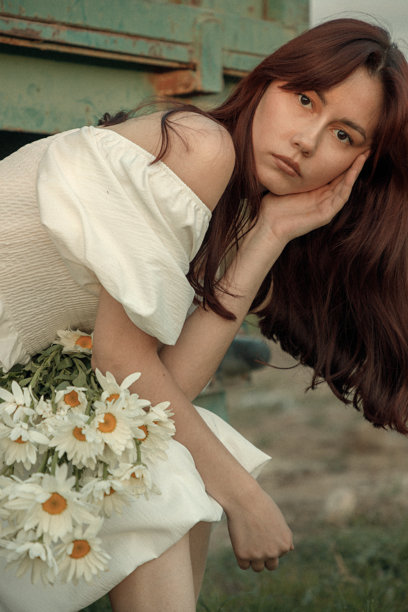 穿着复古风格的年轻女子坐在拖车旁，手中拿着雏菊，画面带有复古颗粒效果。图片下载