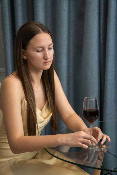年轻女子独自坐在餐桌旁，手持一杯红酒，若有所思地凝视着，反刍着孤独、悲伤和情感动荡的感觉。竖版照片图片下载