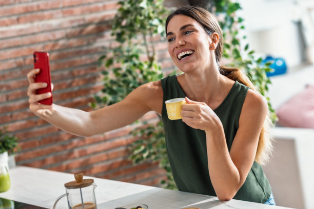 Smiling woman drinking handmade coffee while taking a selfie with her smartphone in the kitchen at home图片下载