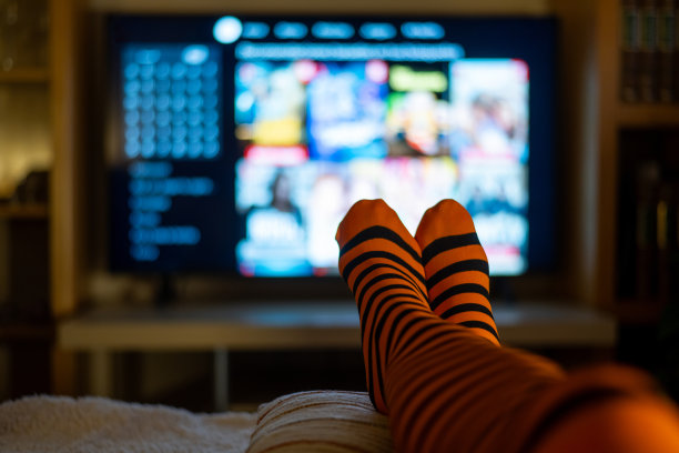 一个人穿着橙黑条纹的袜子，舒适地躺在沙发上，脚搭着，在昏暗的房间里，享受着电视屏幕上播放的恐怖电影。图片下载