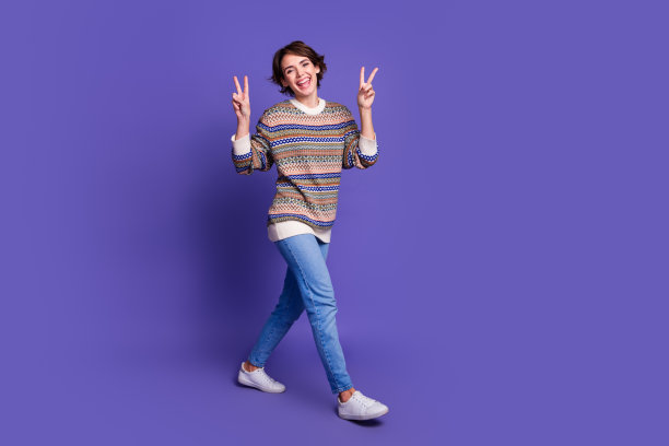 照片：一位穿着时尚服装、竖起胜利手势的可爱、快乐的年轻女子，孤立在紫色背景的空旷空间里行走。图片下载