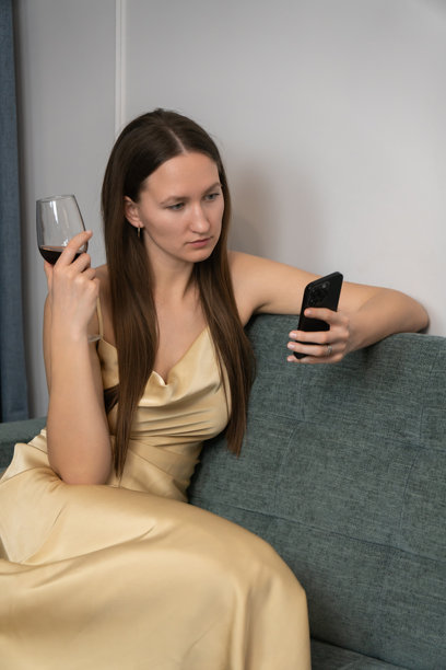 身着优雅连衣裙的年轻女子在家中舒适的沙发上，一边使用智能手机一边享受放松的夜晚，手中还拿着一杯红酒。竖幅照片图片下载