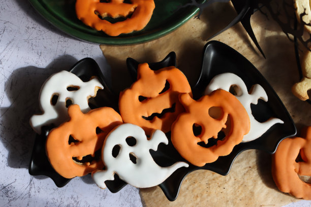 自制万圣节点心的特写图片，包含蝙蝠设计和绿色糖霜饼干，有原味和皇家糖霜两种，还有防油纸、糖霜南瓜灯和鬼魂饼干，以及一个三维纸质树模型，背景为白色和灰色斑驳的纹理，俯视角度。图片下载