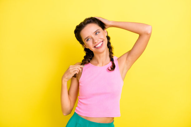 照片：一个可爱快乐的年轻女子，留着夏日发型，梳着辫子，触摸着头发，孤立在明亮的黄色背景上，她望着远方。图片下载
