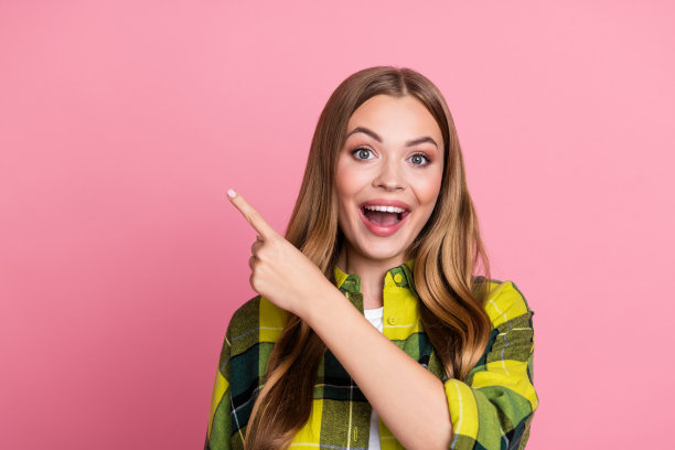 照片肖像，一位有魅力的年轻女子，兴奋地指向空白处，身穿时尚的黄色格子服装，在粉红色背景上孤立。图片下载