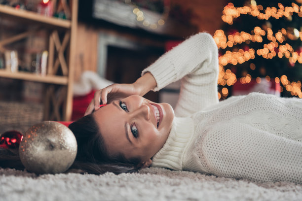 照片：一位可爱的棕色头发女士坐在家里的地毯上，穿着白色毛衣。图片下载