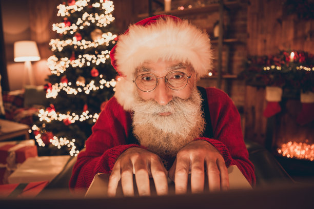 照片：专注的晚归的圣诞老人戴着帽子、穿着套头衫，在北极办公室对着电脑摄像头打字图片下载