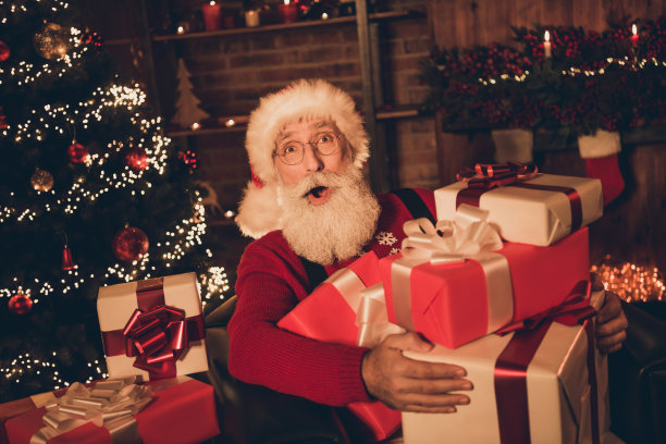 照片：一位穿着圣诞老人服装、面带惊喜表情的滑稽中年男子，在室内家中抱着许多礼品盒。图片下载