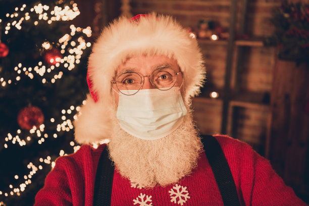 照片：一位戴着医用口罩、采取新冠病毒防护措施的年长圣诞老人，背景是室内的圣诞灯饰。图片下载