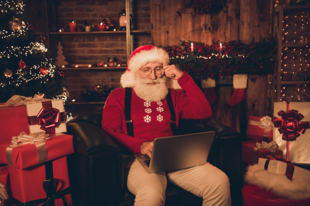 照片：一位戴眼镜、穿着毛衣的圣诞老人坐在装饰的房间里，用笔记本电脑浏览网页。图片下载