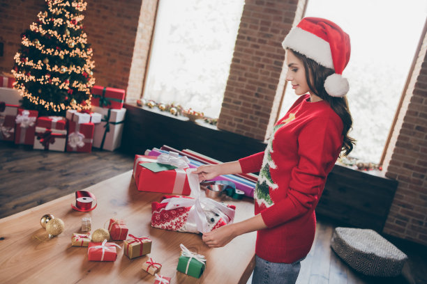 照片：一位穿着印花套头衫、戴着头饰的甜美迷人的年轻女子，正在室内房间、家中打包礼物，脸上带着微笑。图片下载