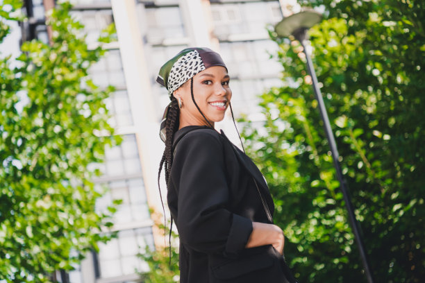 一位自信的年轻女子身穿时尚西装外套，在城市公园里悠闲漫步，背景是绿植和城市建筑，这是一张现代时尚生活方式的拍摄照片。图片下载