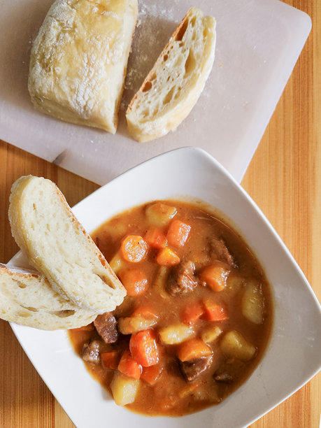 一份令人感到慰藉的牛肉炖菜的图片，里面有软嫩的肉块和蔬菜，旁边配有新鲜的、外脆内软的面包片。图片下载