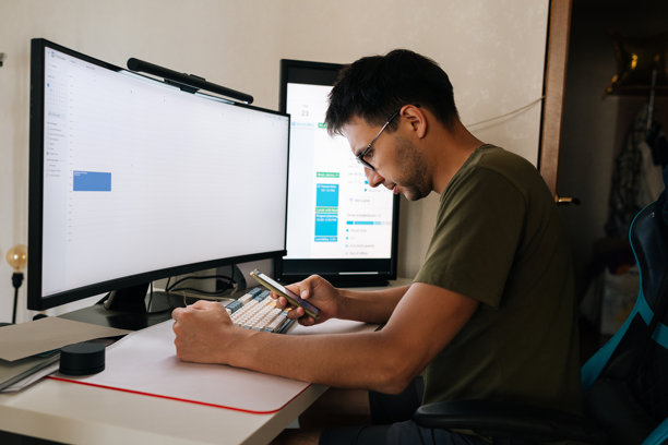 程序员在家办公，坐在书桌前，两台电脑屏幕前，用手机查看日历并安排会议。图片下载