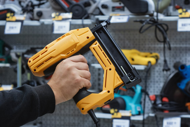 五金店里一个人拿着黄色电动钉枪的特写。用于建筑、木工、家具组装和家庭装修项目的电动工具。图片下载
