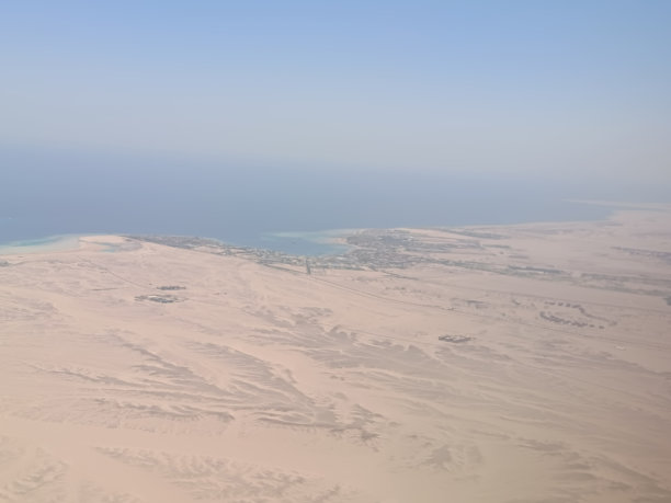 从飞机窗户拍摄的高空照片，展示了广阔的沙漠景观与清澈的蓝色海水交汇，捕捉了沙地的纹理。图片下载