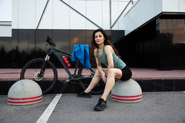 年轻健美的女性穿着运动服，在街上与她的黑色山地自行车坐在一起，在夏日阳光下休息。户外运动锻炼。图片下载