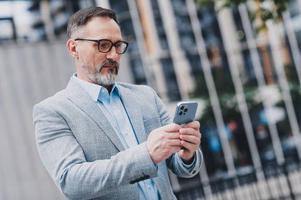 商务人士在现代都市街头，身着西装，戴着眼镜，使用智能手机的素材照片，适合商务专业人士。图片下载