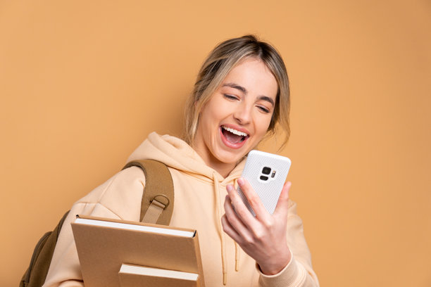 身穿米色衣服的快乐高加索女性正在用手机观看视频和照片，与上学、教育、学生概念相关。图片下载