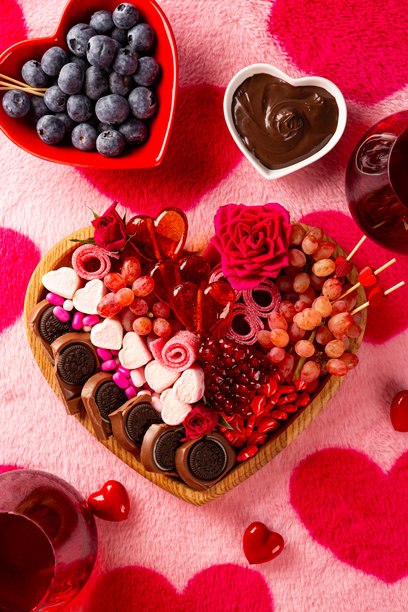 心形零食拼盘，内含浆果、巧克力和糖果，为情人节准备，配有两杯红酒，背景为粉色带爱心图案的竖幅照片。图片下载