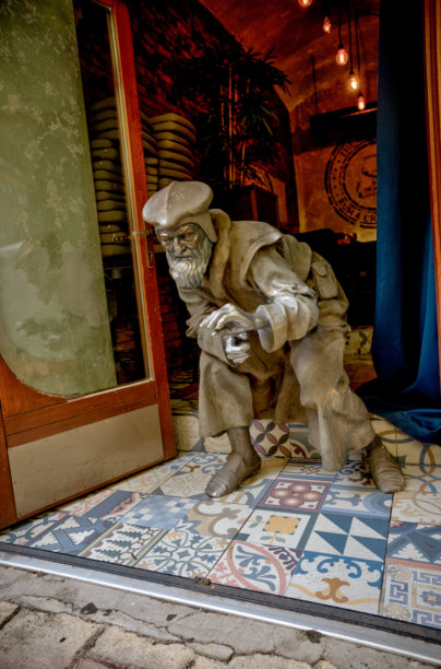 一个真人大小的驼背中世纪人物雕塑在当地一家商店的入口处迎接访客，它矗立在色彩鲜艳的图案瓷砖上。艺术家和创作日期不详。图片下载