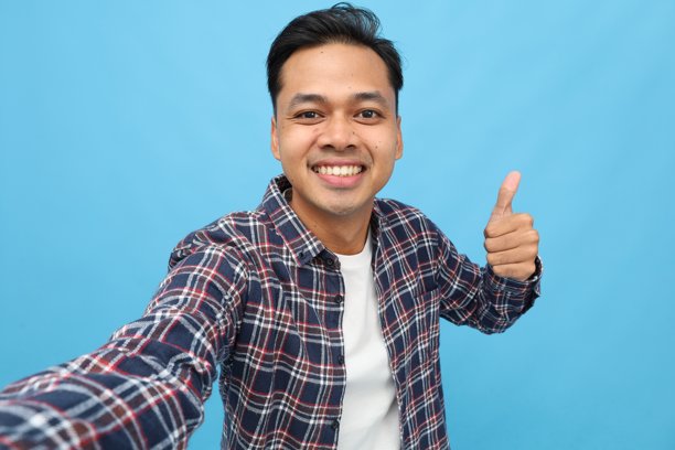 年轻成功的亚洲男性，穿着休闲服装，用手机拍摄自拍视角照片，竖起大拇指，背景为蓝色，独立。图片下载