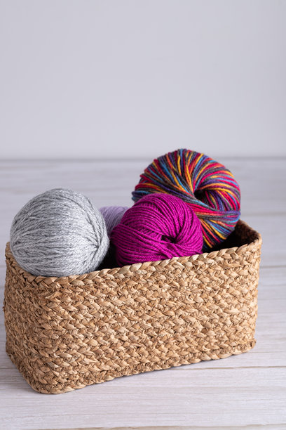 浅色木桌上的柳条篮，里面装着灰色、紫红色和彩色的毛线球。垂直的极简主义照片，带有中性背景和留白。编织毛线和创意生活方式的概念。图片下载