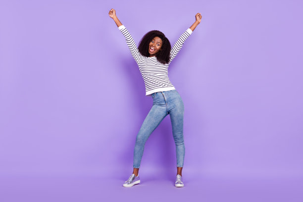 一名年轻女子双手握拳，身穿条纹套头衫和牛仔裤，身处紫色背景前，全尺寸照片。图片下载