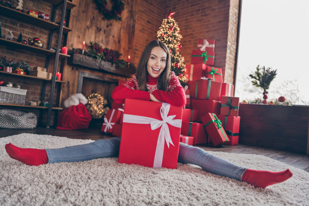照片：一位穿着红色套头衫、面带笑容、手持圣诞礼物的迷人年轻女子，在室内的家中，面露兴奋之色。图片下载