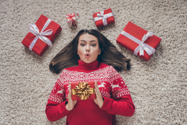 照片：一位震惊又印象深刻的年轻女子，穿着红色套头衫，在室内房屋房间里拿着圣诞礼物。图片下载