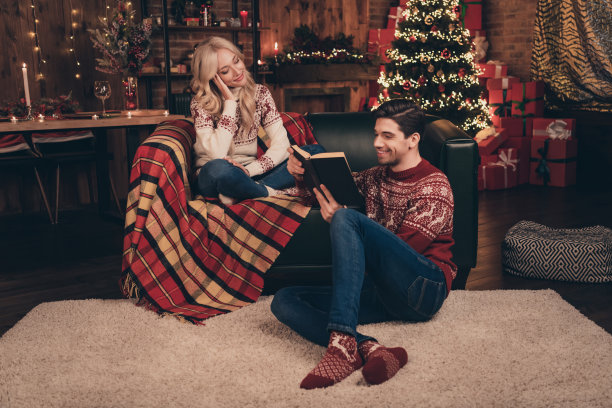 照片：一对漂亮的、积极的夫妇，戴着印花毛衣，在室内（房子、家、房间）微笑着阅读和聆听新年童话故事。图片下载