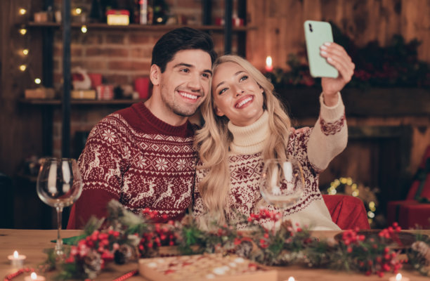 照片：一对 pretty 迷人的夫妇，穿着印花套头衫，戴着现代设备，在室内房间、家中享受圣诞节，进行视频通话，脸上带着笑容。图片下载