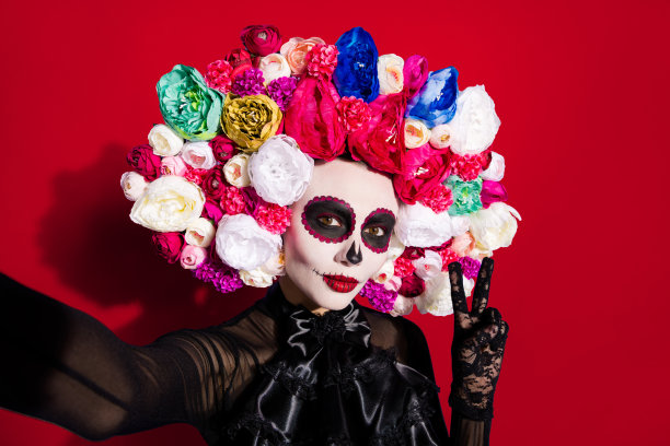 特写照片，展示一位身着花卉头饰服装的传统拉丁民间传说女巫，在亡灵节（Day of the Dead）期间化着诡异吓人的妆容，戴着假面具，在孤立的紫色背景前自拍，并摆出V字手势。图片下载