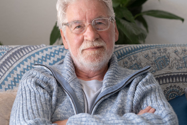 一位慈祥的留胡子老人，双臂交叉，在家里的沙发上放松，面带微笑看向镜头。一位享受退休生活的年长帅气男子的头像。图片下载