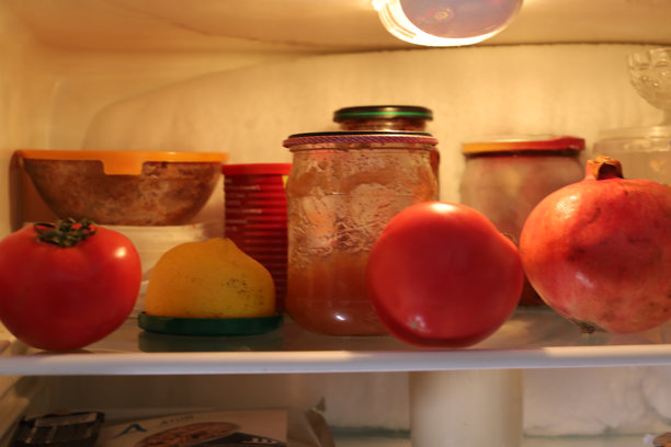 冰箱内玻璃架上的食物，包括鲜亮的番茄、柠檬和石榴，旁边是罐子里自制的果酱。这张特写照片展示了厨房电器内的食物储存和储备。图片下载